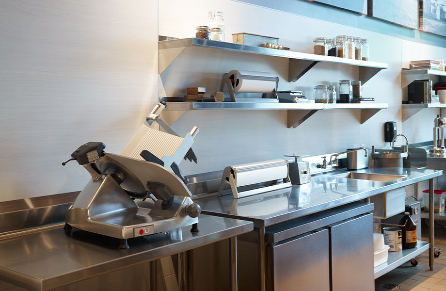 An industrial kitchen with stainless steel countertops, shelving, a meat slicer and food preparation tools